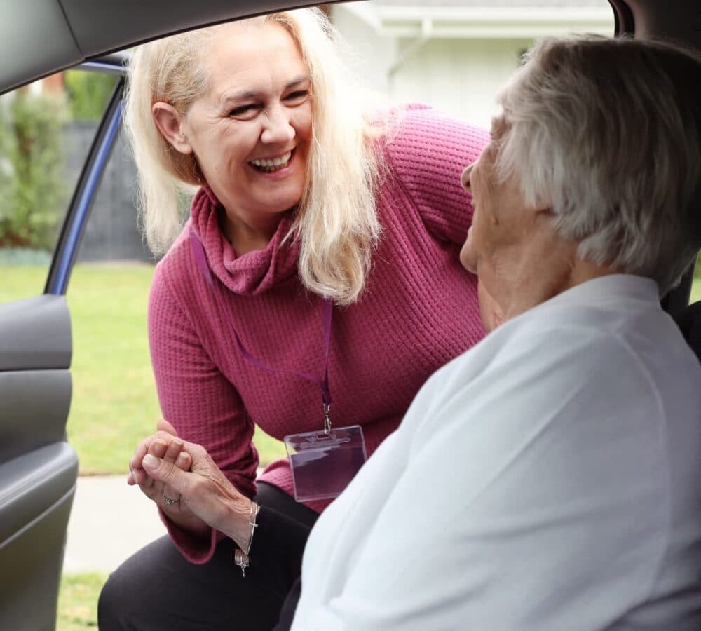 Senior woman going out of the car being helped by her carer smiling and wearing pink top