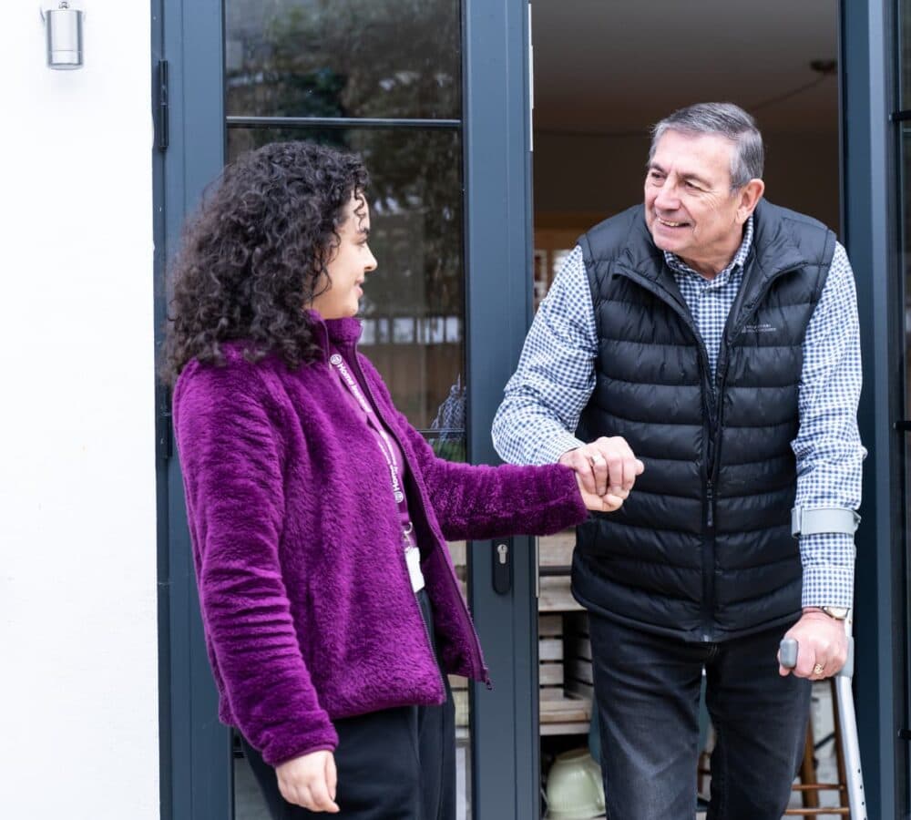 Tall senior man going out the door using a crutch while being assisted by his carer wearing purple and with curly hair
