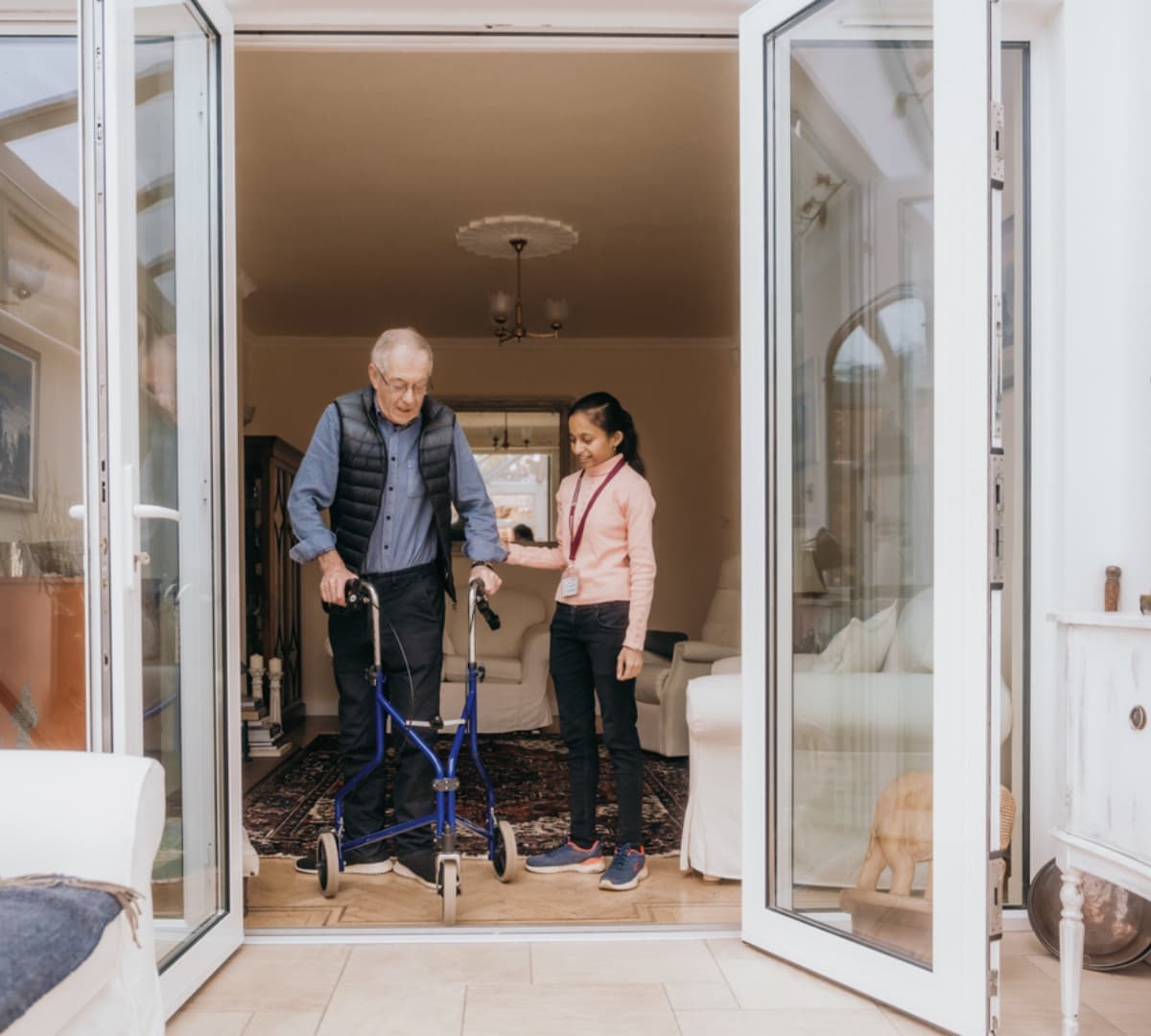 A senior man with grey hair walking and going outside the house while using a walker with his carer with long black hair guiding and helping him.