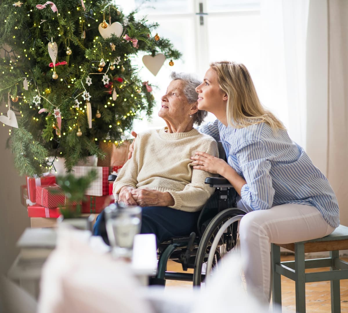 Senior woman on a wheelchair with her daughter sitting behind her inside their house and looking at the window with Christmas tree beside her
