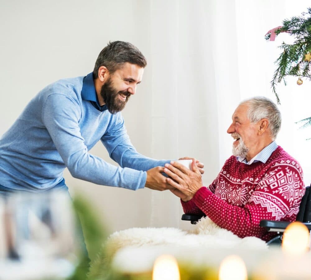 A senior man sitting on a wheelchair and wearing red sweatshirt while receiving gift from his son inside their home with Christmas decorations