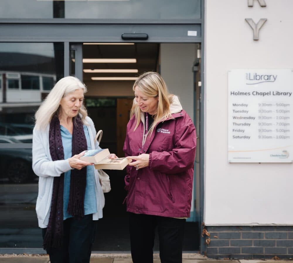 Senior woman with white hair wearing sweaters and walking outside the library while holding a book with her carer