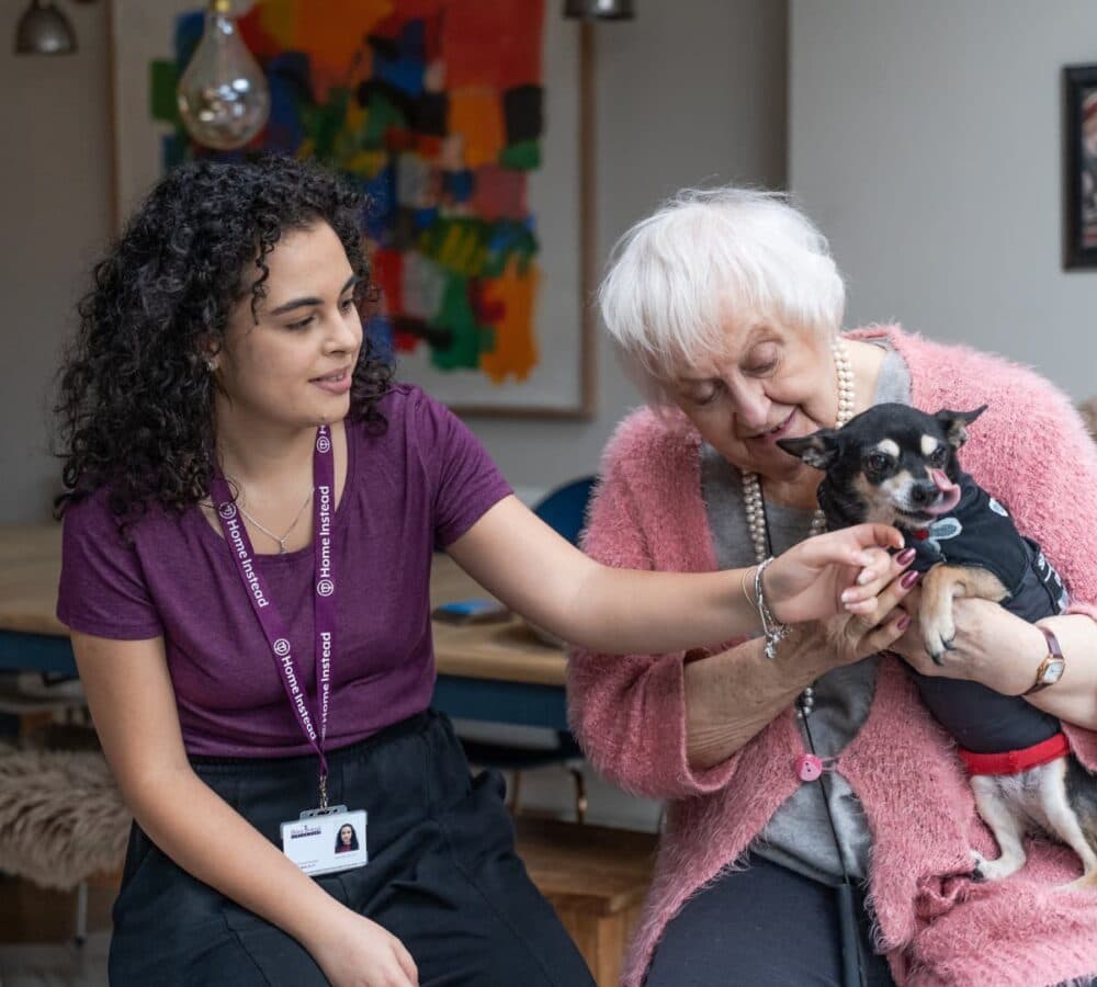 Senior woman wearing pink and with white hair holding a dog with her carer wearing purple and with curly hair both smiling and inside the house