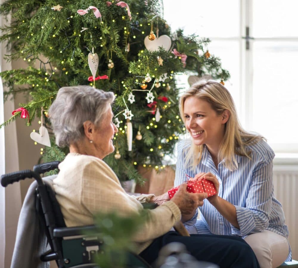 Senior woman sitting on a wheelchair while receiving a gift from her younger daughter wearing blue and sitting on a couch with Christmas tree on the background