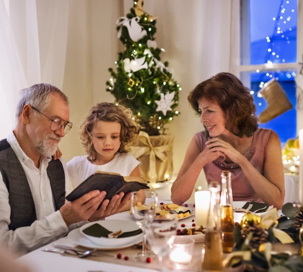 Senior man holding a book with granddaughtr and his daughter sitting with him on a table with Christmas tree on the background