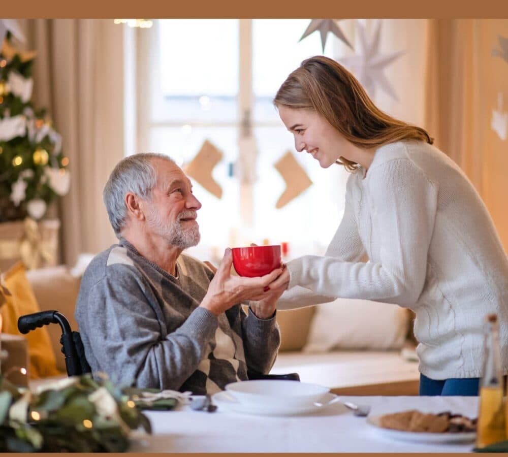 Senior man with grey hair and sitting on a wheelchair holding a red mug from his daughter with Christmas tree and decors in the background