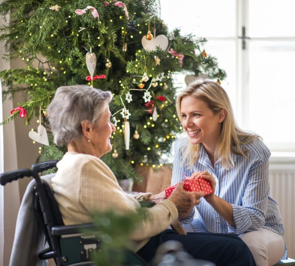 Senior woman with daughter giving her a gift while sitting on a chair and with a Christmas tree beside her