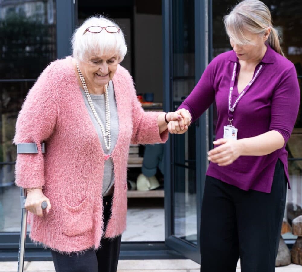 Senior woman walking out the door with white hair and pink sweater and with her carer helping her while using a crutch