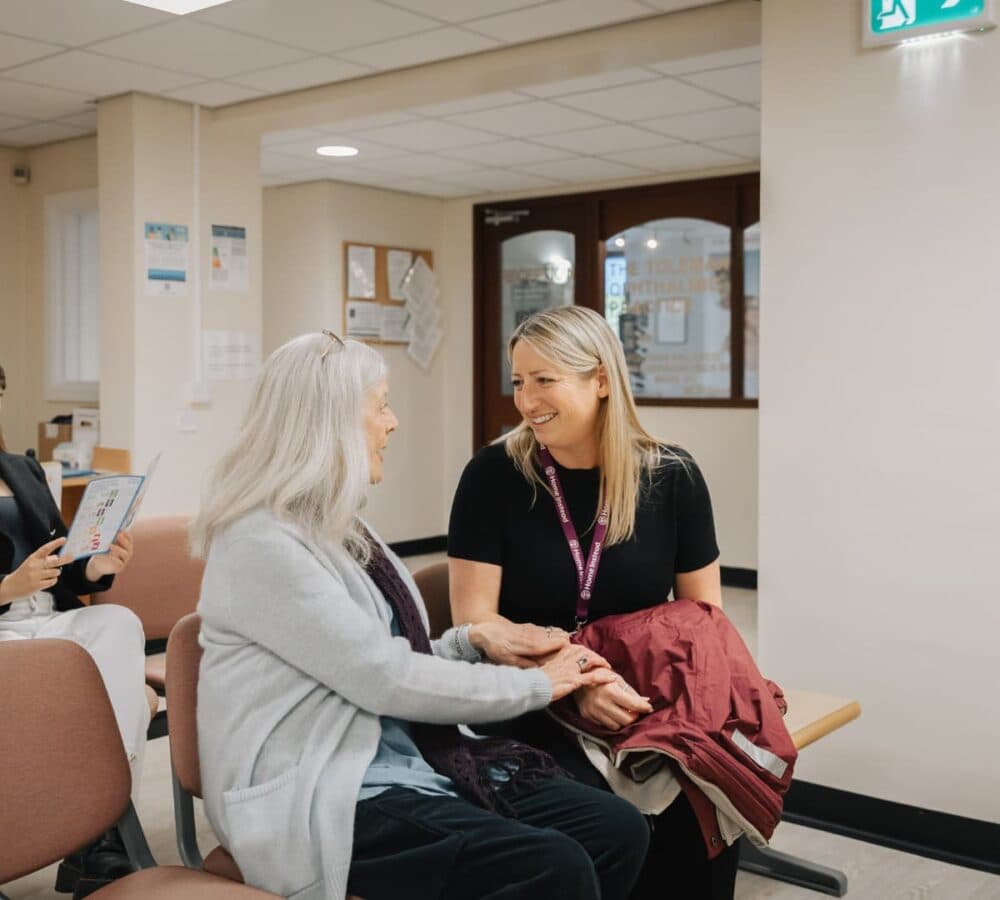 A senior woman and a staff member smiling and talking in a clinic waiting area. - Home Instead