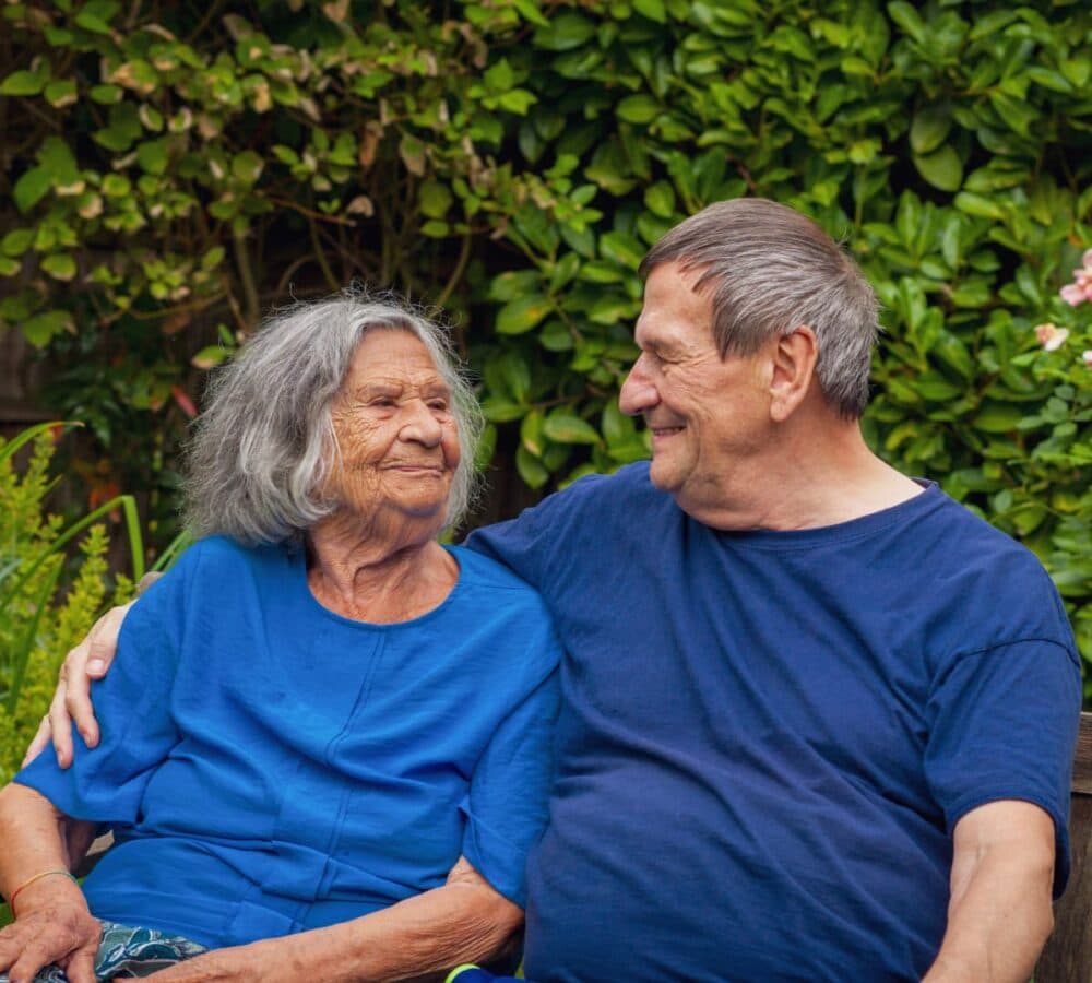 Senior woman sitting on a bench wearing blue and with grey hair looking at her son and smiling in the garden