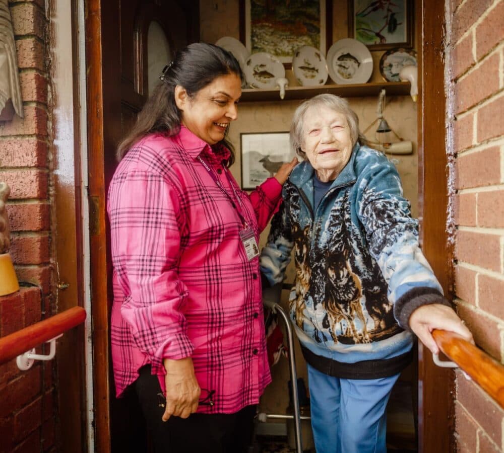 Senior woman standing with white hair and being assisted by her carer inside her home