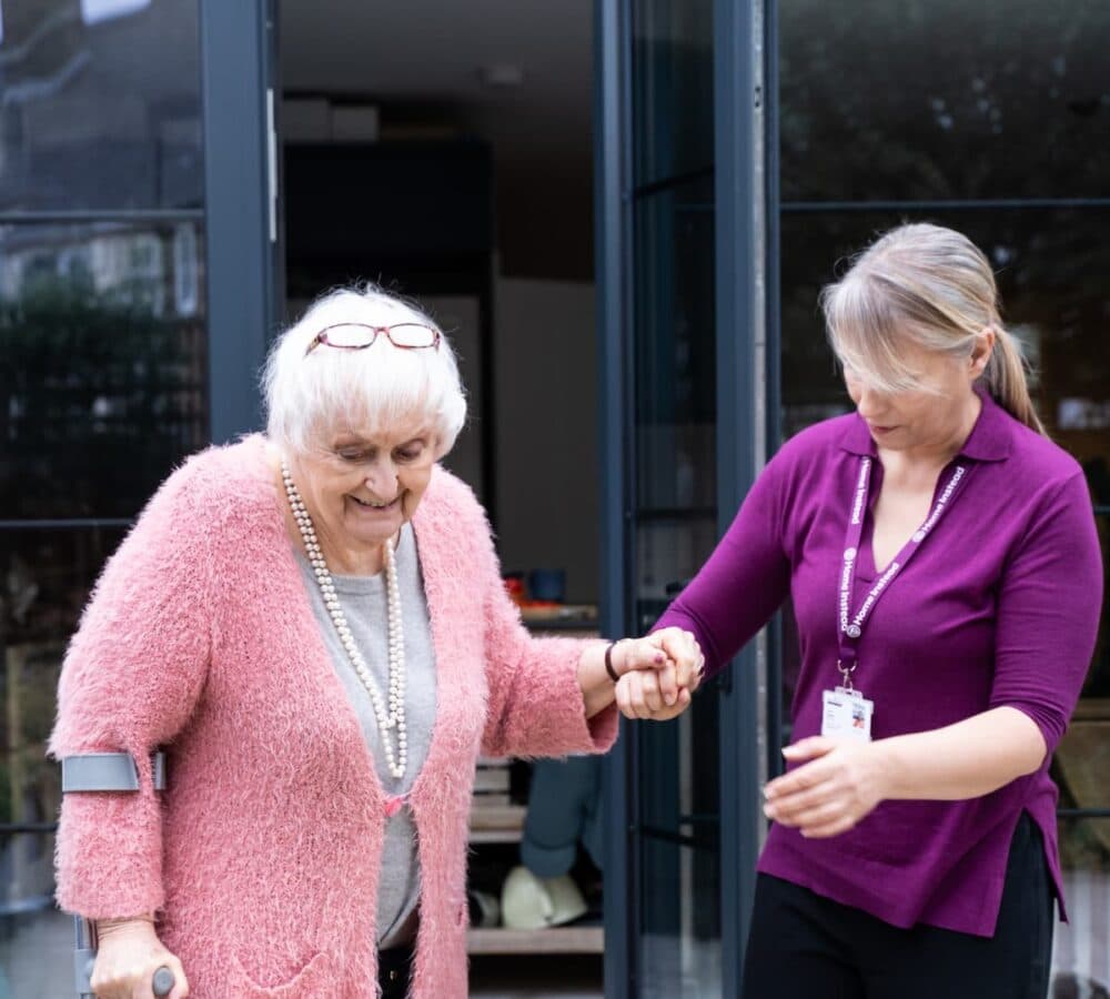 Senior woman with white hair and wearing pink top with her carer going out of the door