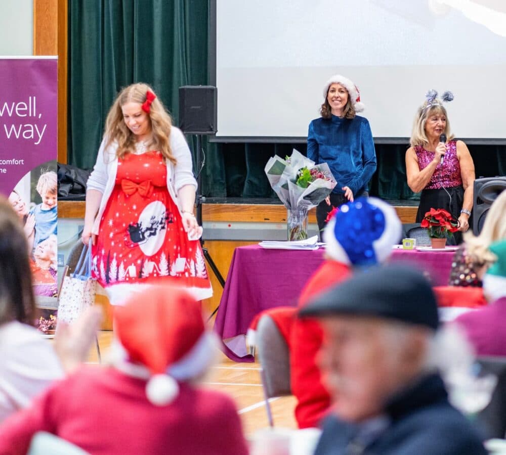 Two women hosting wearing blue with santa hat and another older lady wearing shiny pink top and holding a microphone