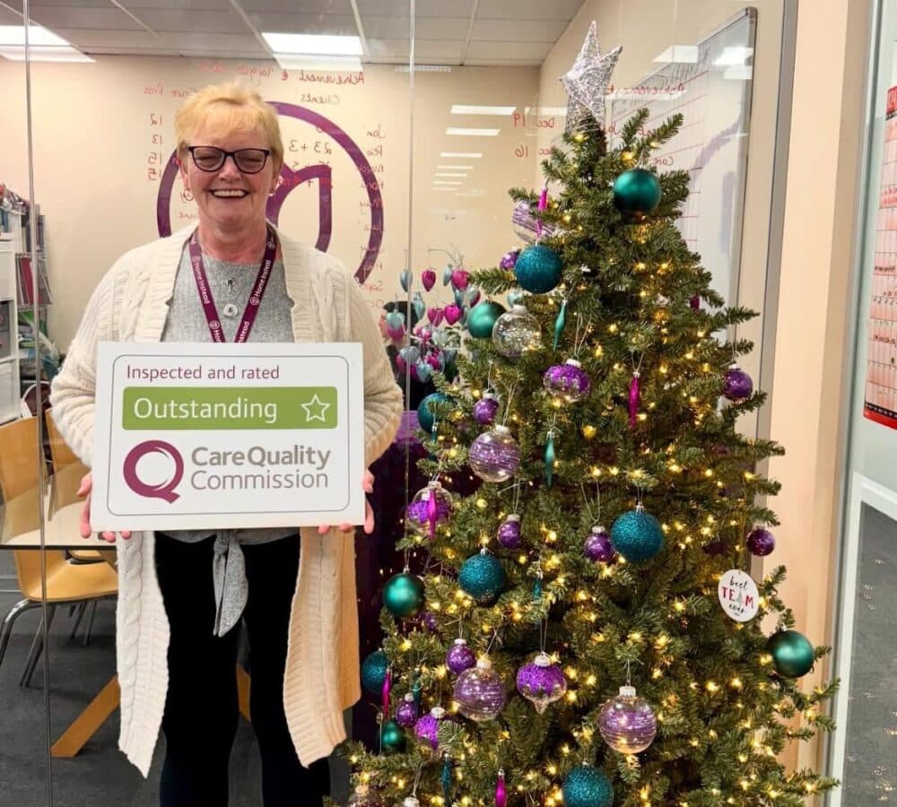 Woman with short hair and wearing eyeglasses standing and smiling beside a christmas tree while holding a CQC award sign inside the office
