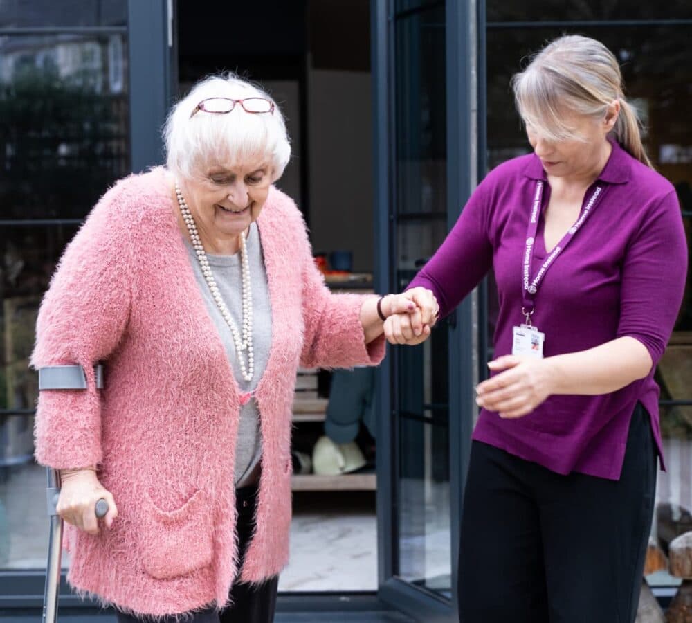 Woman helping a senior woman wearing pink with grey hair while using crutches while walking
