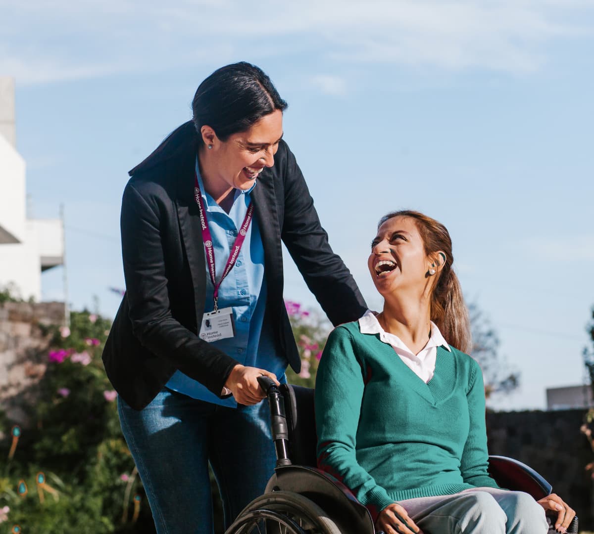 Woman on a wheelchair being helped by her carer with black hair outdoors with a clear blue sky and plants at the background