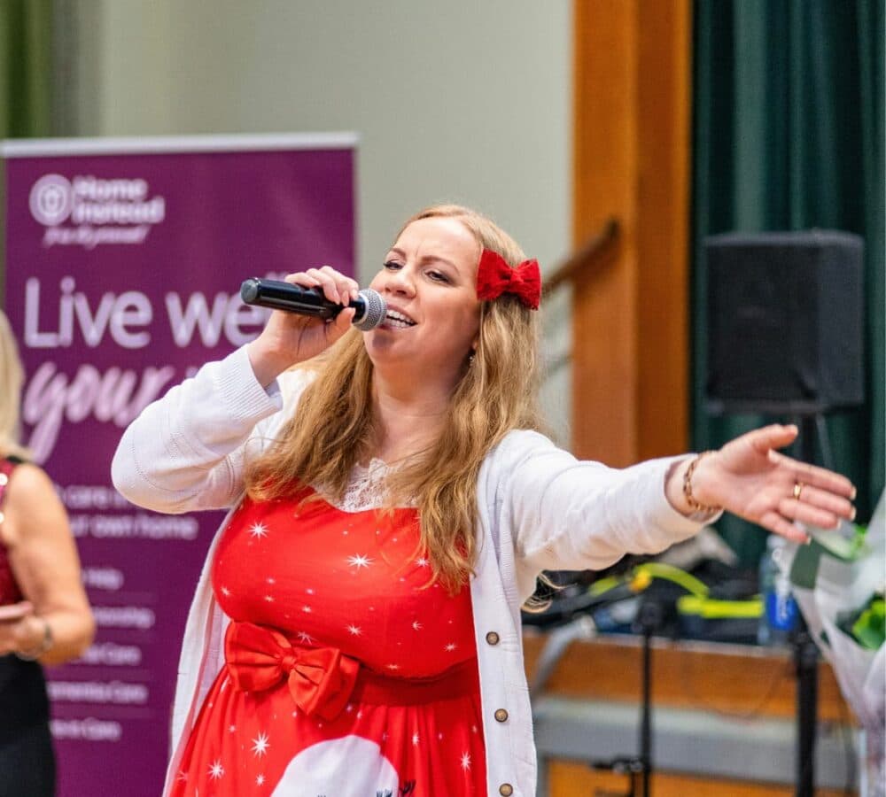 Woman with long blonde hair and with a red ribbon on her hair wearing a red dress singing inside the hall