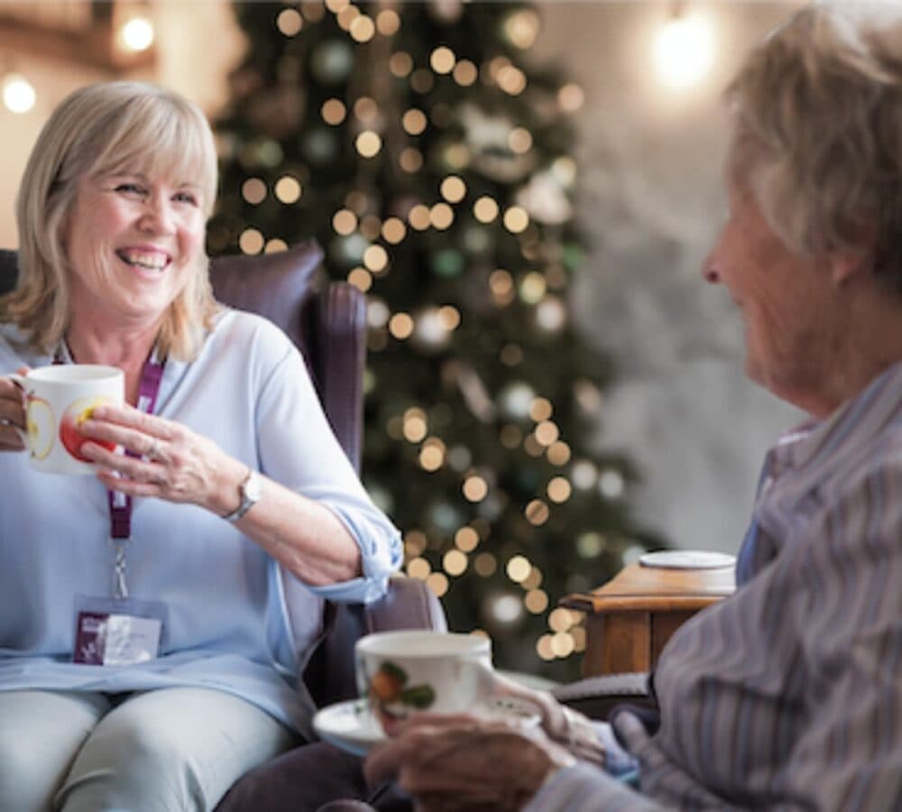 Woman with a senior both smiling while having a cup of tea and with a Christmas tree at the background