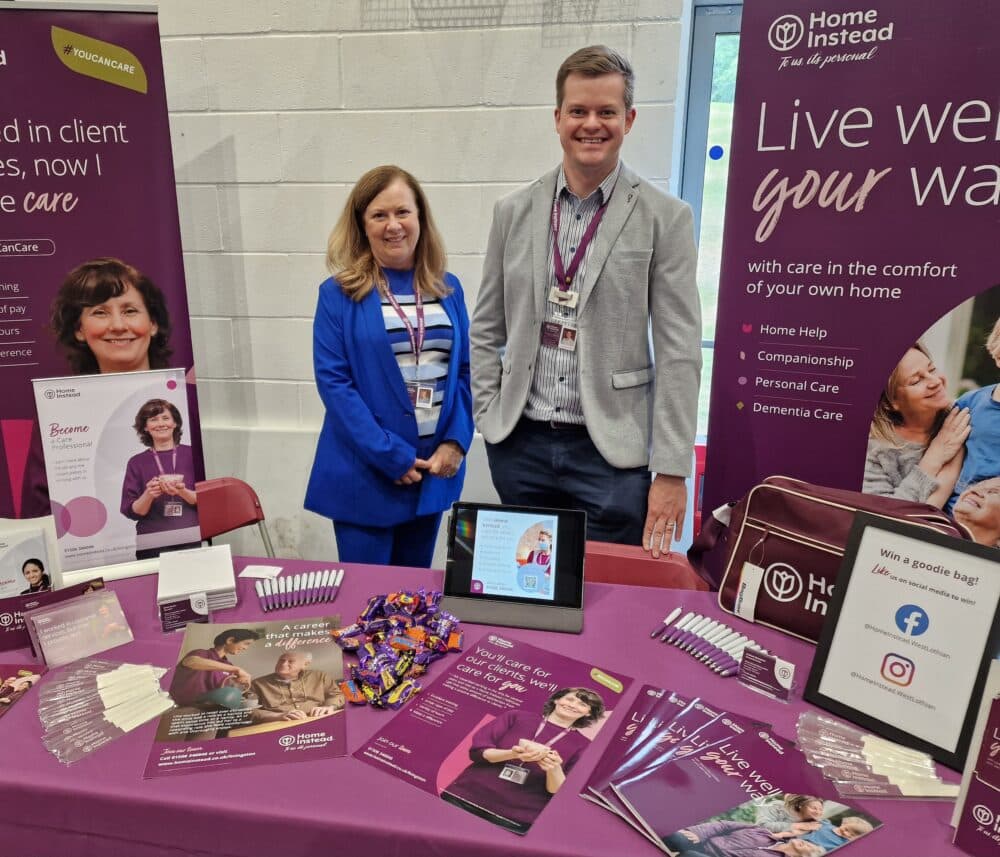 Two people standing at event stall with branded literature and posters - Home Instead