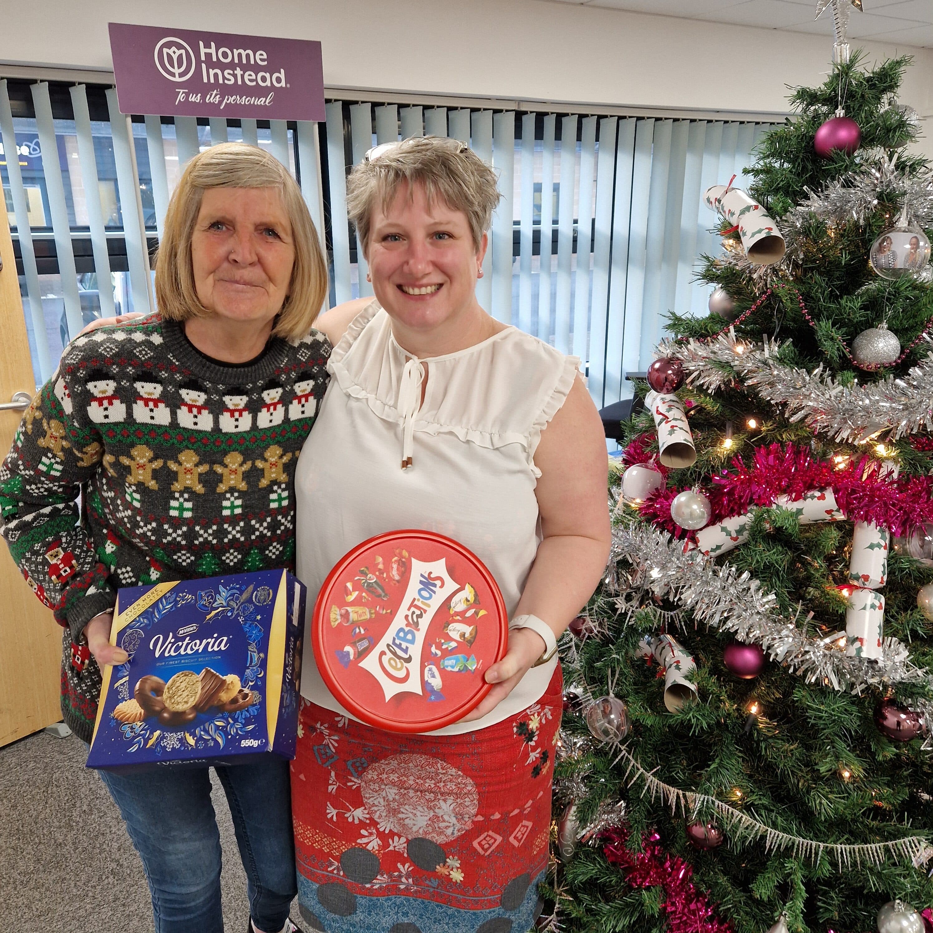Two women smiling by a Christmas tree, holding biscuit tins, with a "Home Instead" sign in the background. - Home Instead