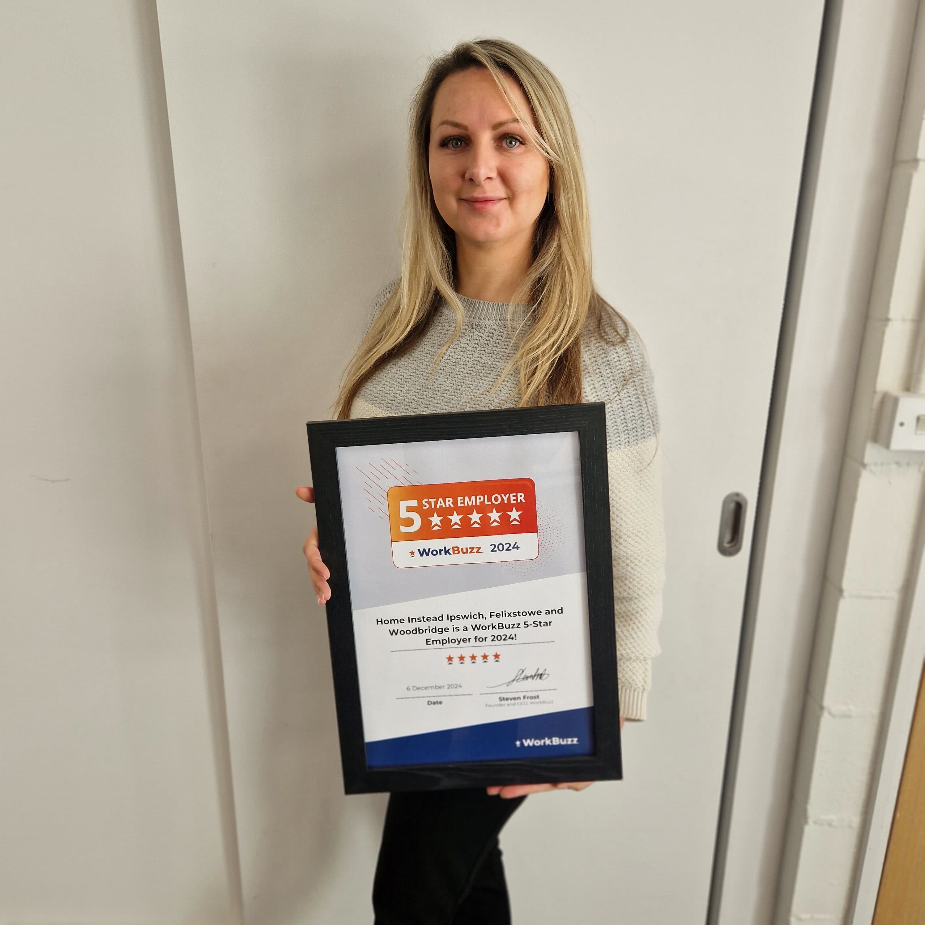 A woman holding a framed 5 Star Employer 2024 award, standing against a light-colored wall. - Home Instead