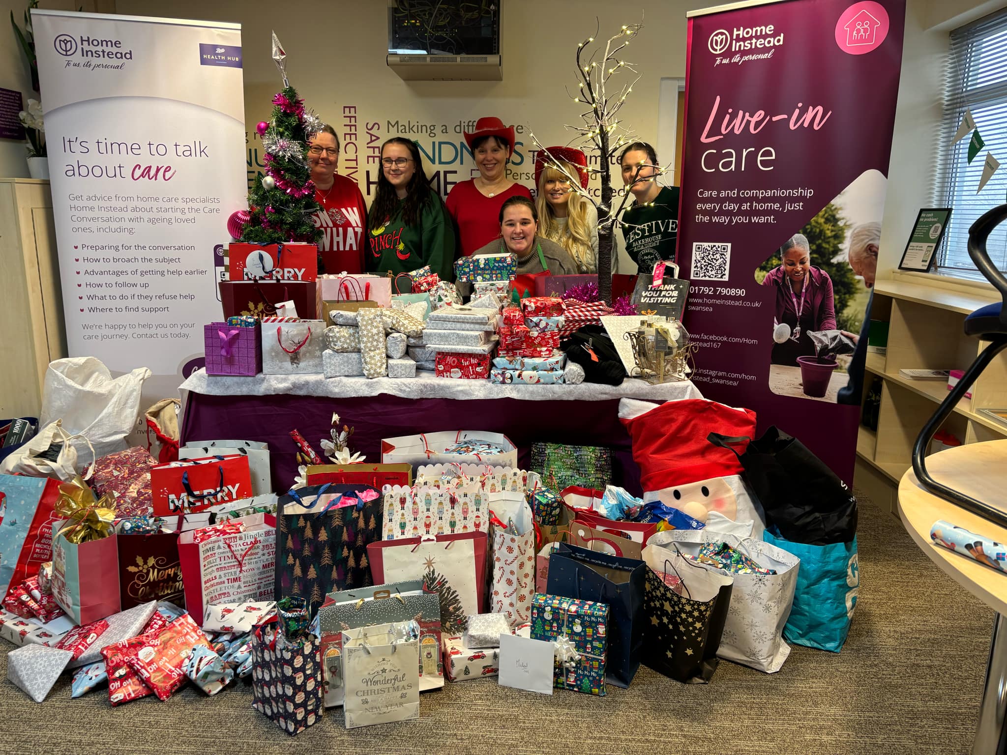 6 women posting alongside Home Instead promotional banners alongside hundreds of wrapped Christmas gifts