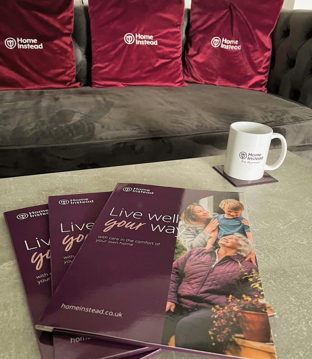 Three Home Instead brochures and a mug on a table with maroon Home Instead cushions on a grey sofa behind. - Home Instead