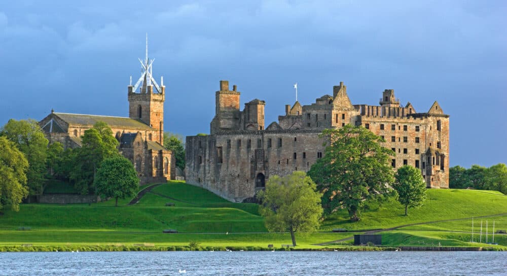 Medieval castle ruins and church on a green hill by a river under a dramatic, cloudy sky. - Home Instead