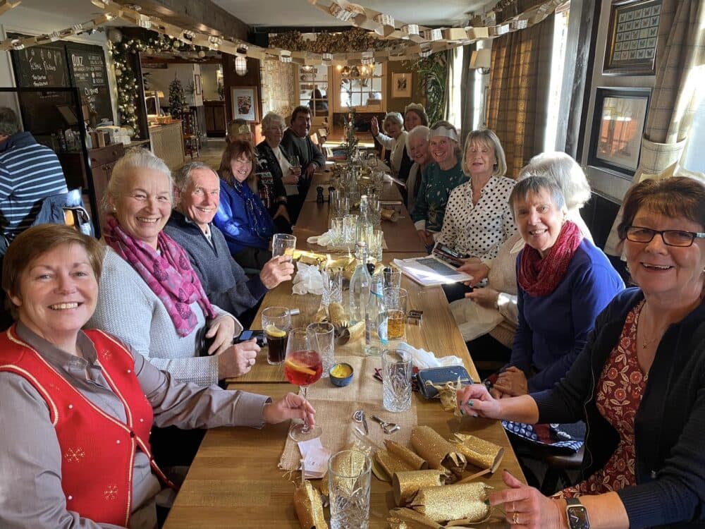 A group of older adults smile and enjoy drinks together around a long wooden table in a cozy, well-lit room. - Home Instead