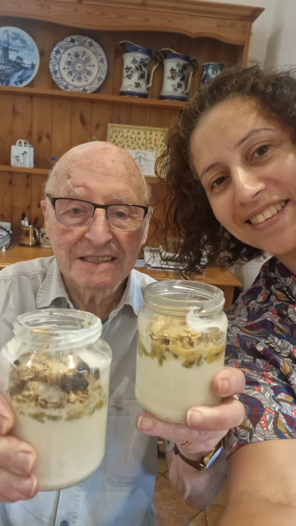 Smiling man and woman hold up jars of parfait or yogurt, posing together in a cozy kitchen. - Home Instead