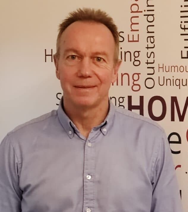 Man in a light blue button-up shirt stands in front of a wall with motivational words in various fonts. - Home Instead