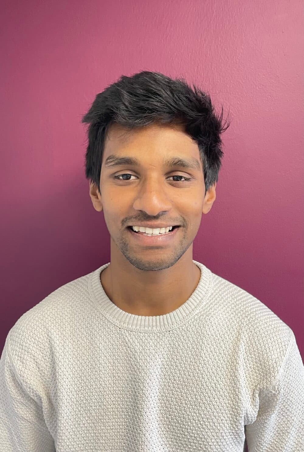 Smiling man with dark hair in a white sweater, standing in front of a magenta wall. - Home Instead