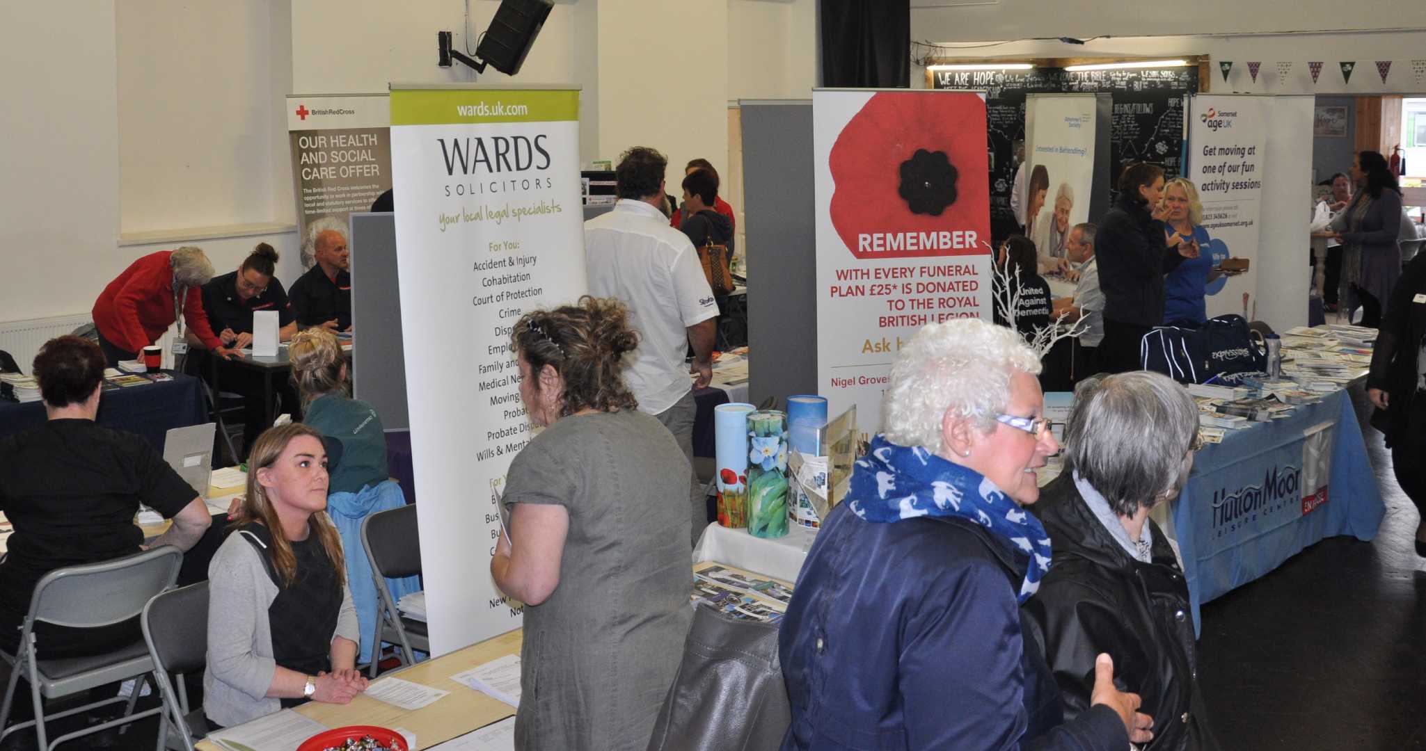 People visit various informational booths at an indoor community event or fair, engaging with exhibitors. - Home Instead