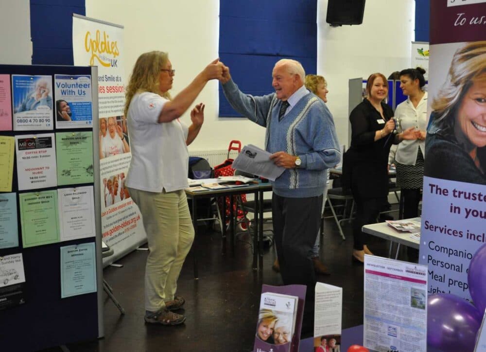 An elderly man and woman smile and dance together at a community event with informational booths. - Home Instead