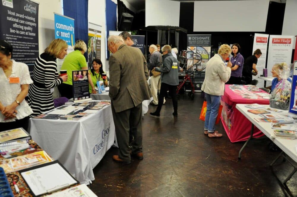 People visiting information booths at an indoor community event or fair, talking with stall representatives. - Home Instead