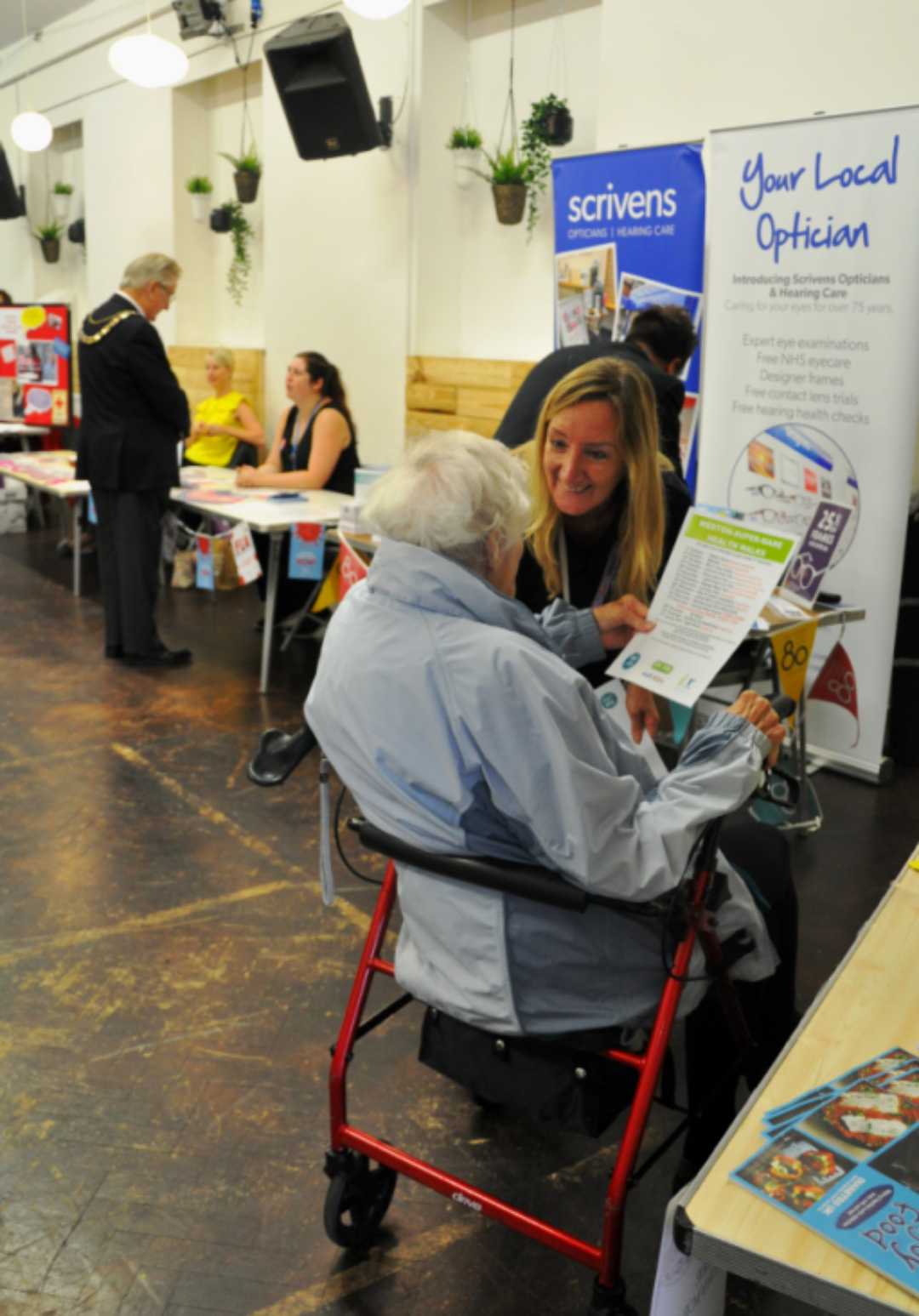 A woman speaks to an elderly person in a walker at an indoor community health event with information tables. - Home Instead