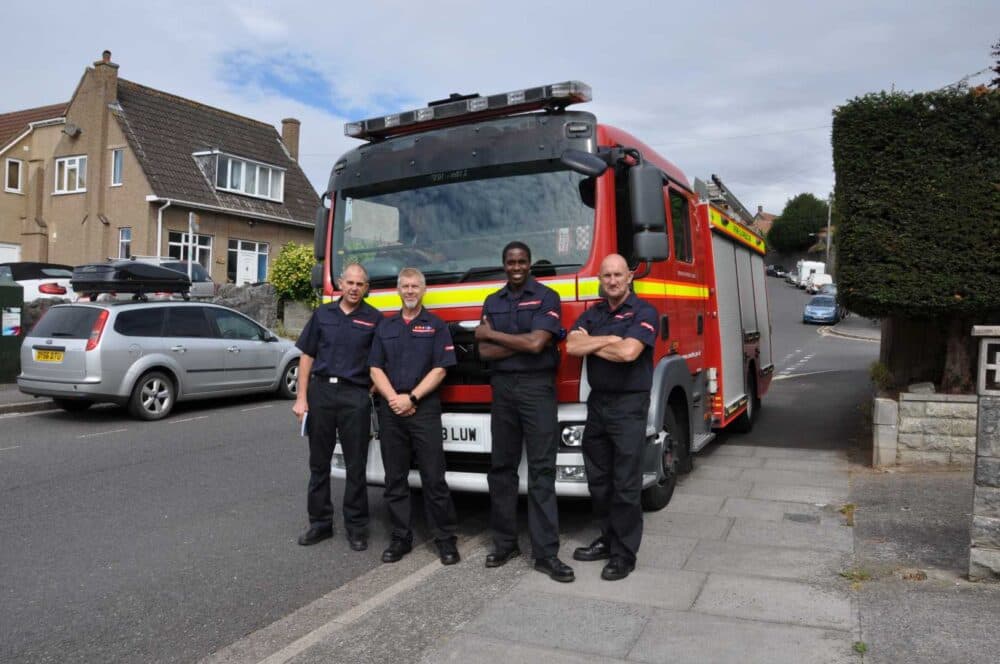 Four firefighters stand in front of a fire engine parked on a residential street. - Home Instead