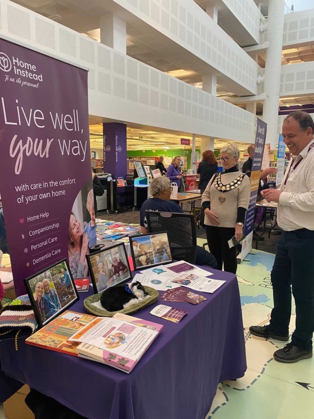 People at an indoor care services fair stand next to a "Live well, your way" Home Instead banner and table display. - Home Instead