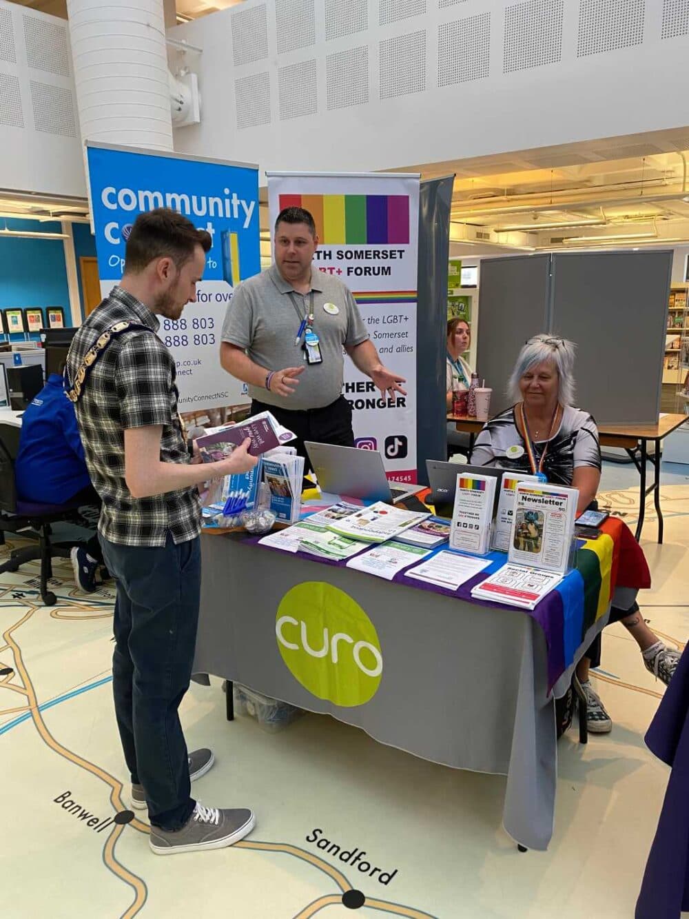 Three people talk at an information booth for Curo and the LGBT+ Forum at a community event indoors. - Home Instead