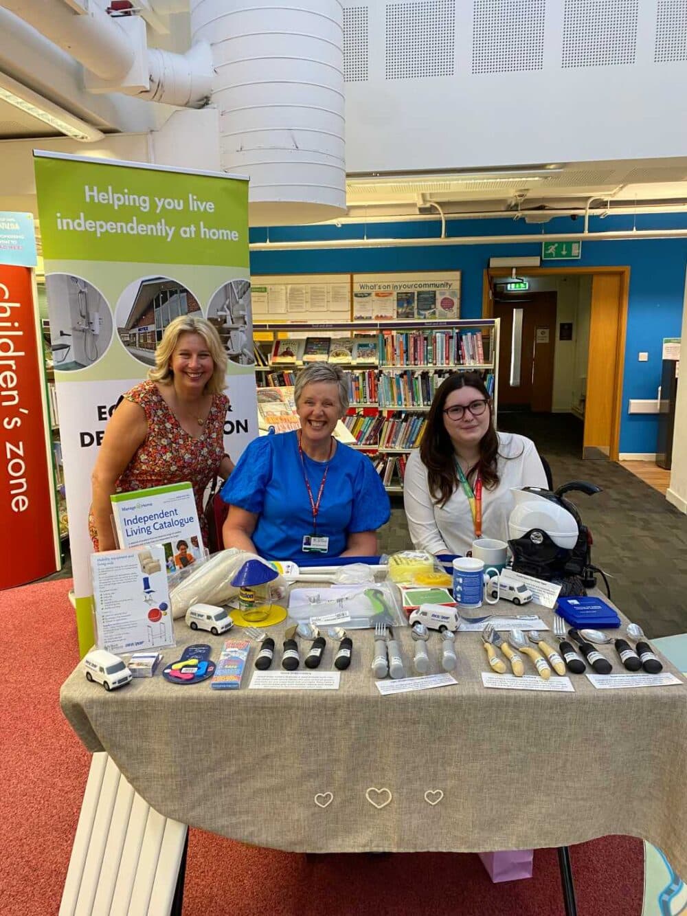 Three women at a display table with assistive products in a library, promoting independent living. - Home Instead