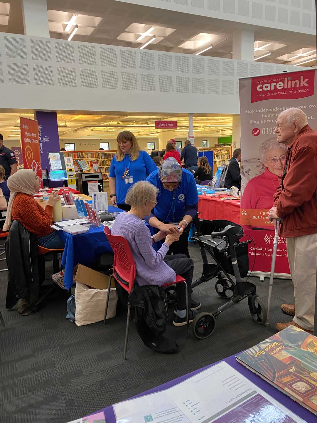 A nurse helps an elderly woman at a community health fair in a library, with others at nearby tables. - Home Instead