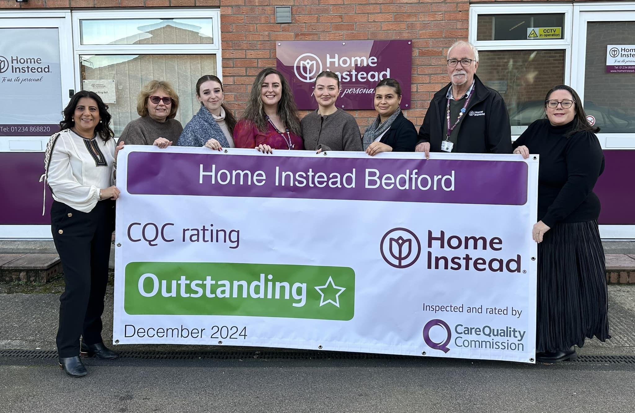 Eight people smile holding a Home Instead Bedford banner showing an "Outstanding" CQC rating for December 2024. - Home Instead