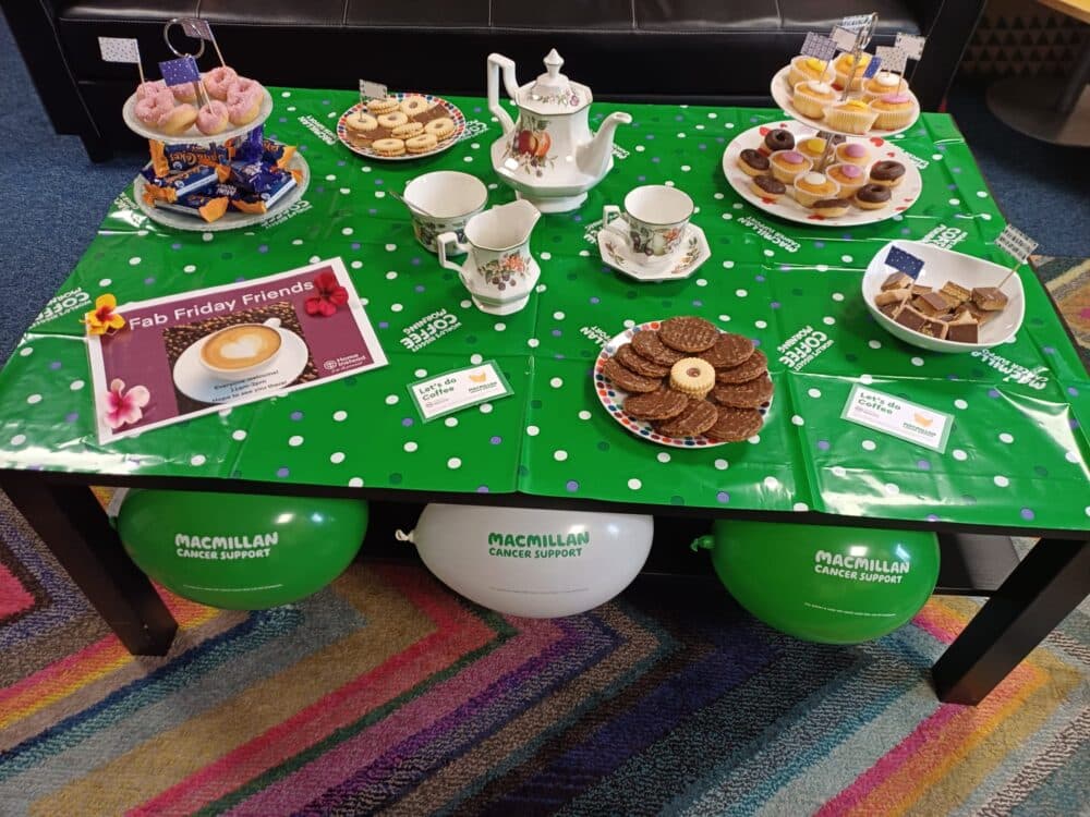 A table with tea, cakes, and treats on a green Macmillan Cancer Support tablecloth, with balloons underneath. - Home Instead