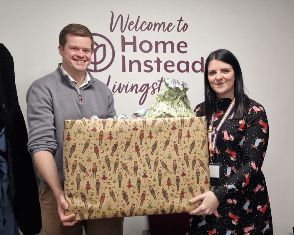 Two people holding a box of Christmas presents in front of a sign - Home Instead