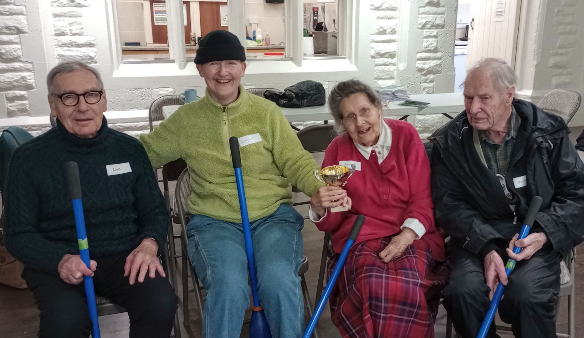 Four elderly people sitting indoors, smiling, holding blue sticks; one woman holds a trophy. - Home Instead