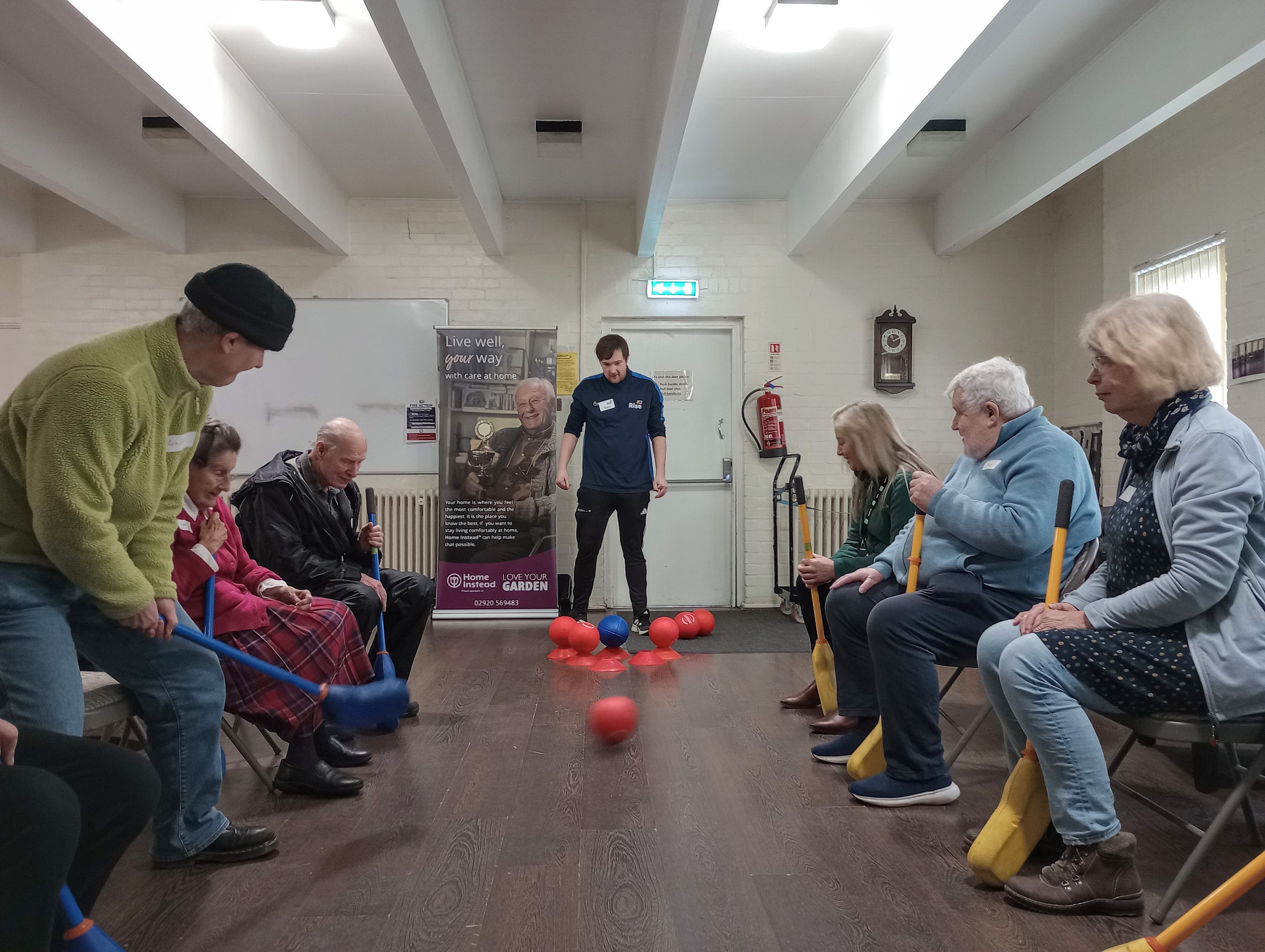 A group of seniors sit in a circle playing a seated game with plastic bats and red balls indoors. - Home Instead