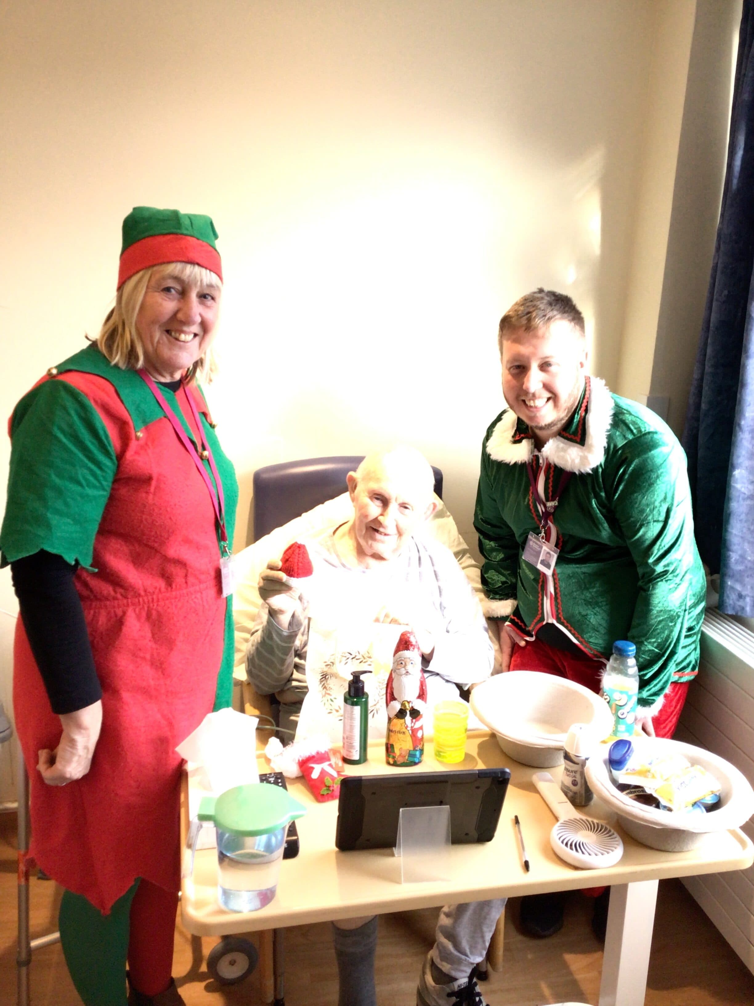 Two people in elf costumes visit a smiling elderly man in a hospital room with Christmas treats on the table. - Home Instead
