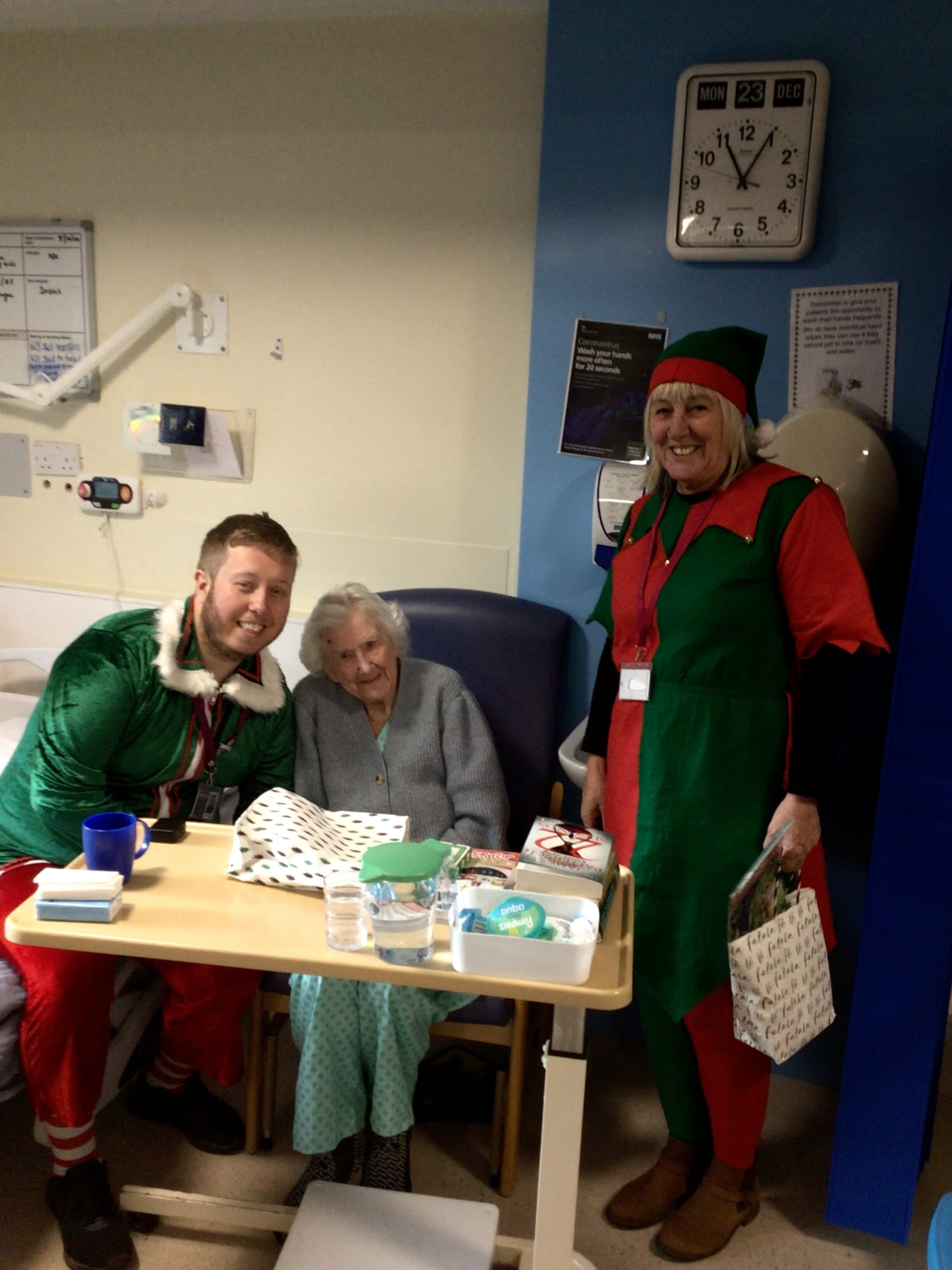 Two people in elf costumes visit an elderly woman in a hospital room, smiling beside her bed. - Home Instead