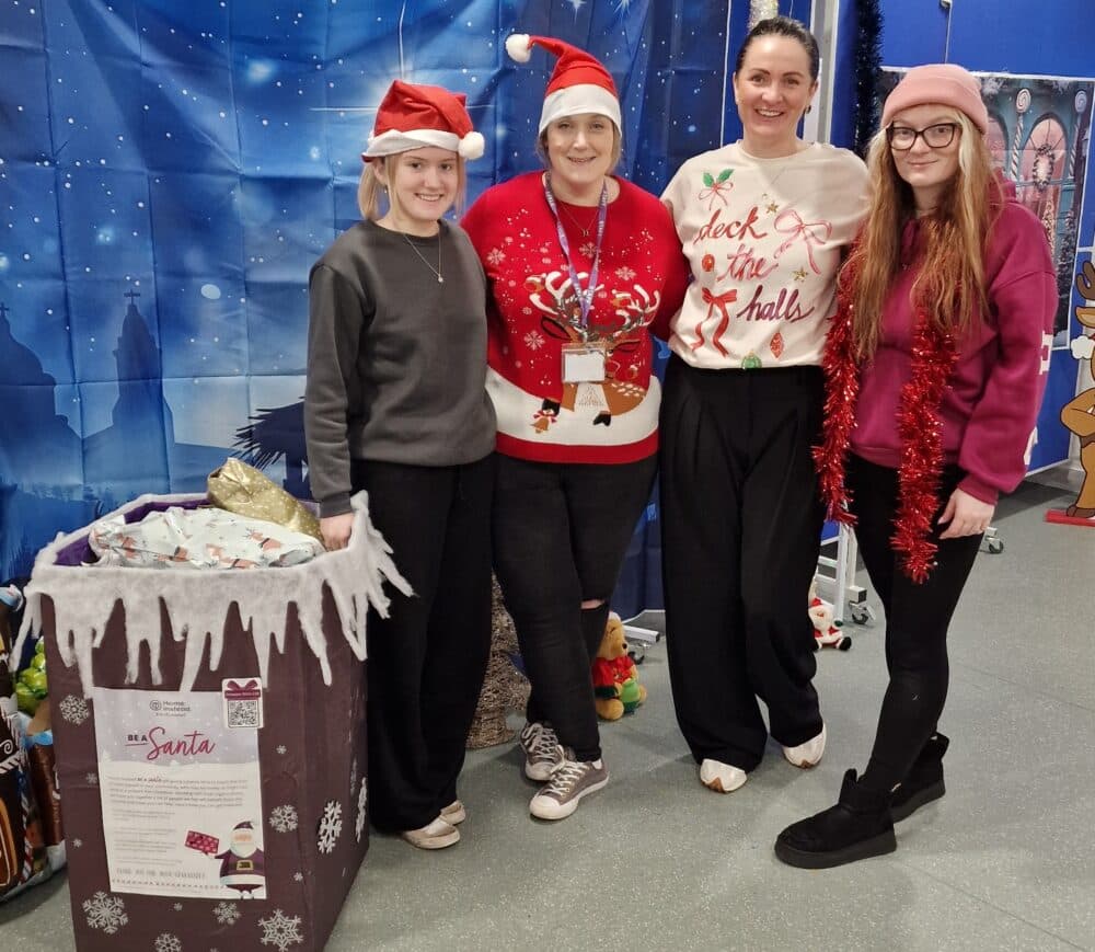 Four people wearing Christmas hats and jumpers, standing next to a collection bin - Home Instead