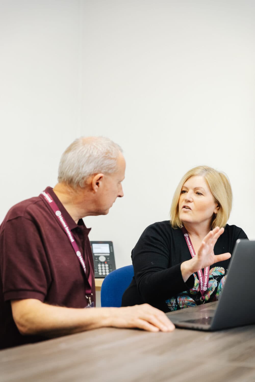Two colleagues sit at a table, having a discussion while looking at a laptop. - Home Instead